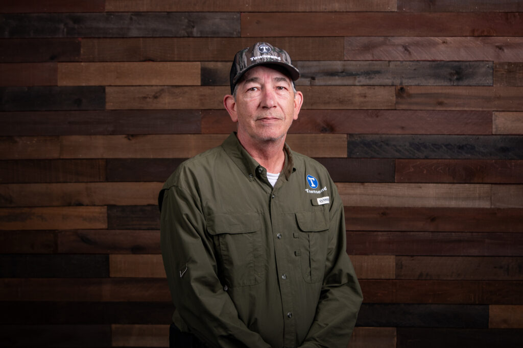 man with a dark green button up shit and a camo hat in front of a wooden background