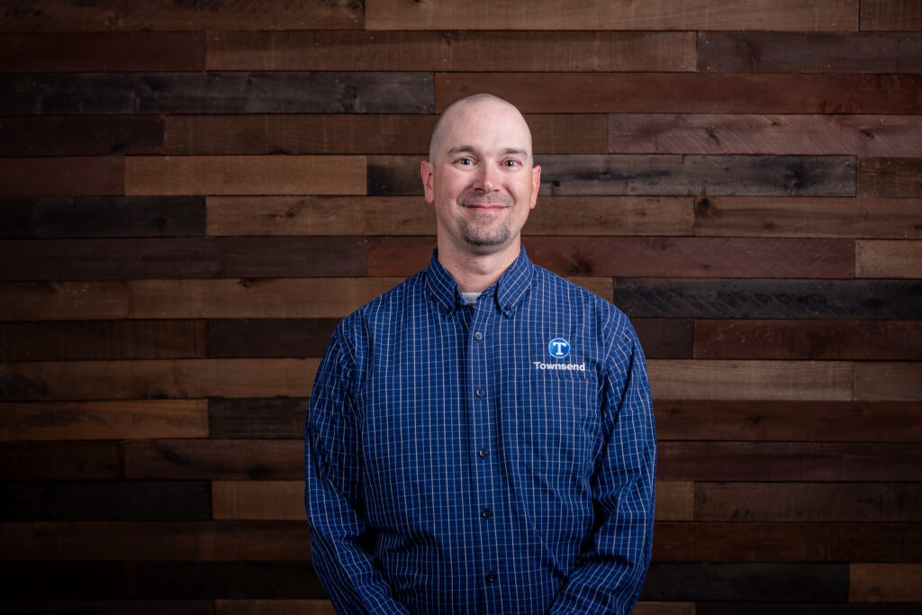 bald man with a beard in a blue and white striped shirt standing in front of a wooden background