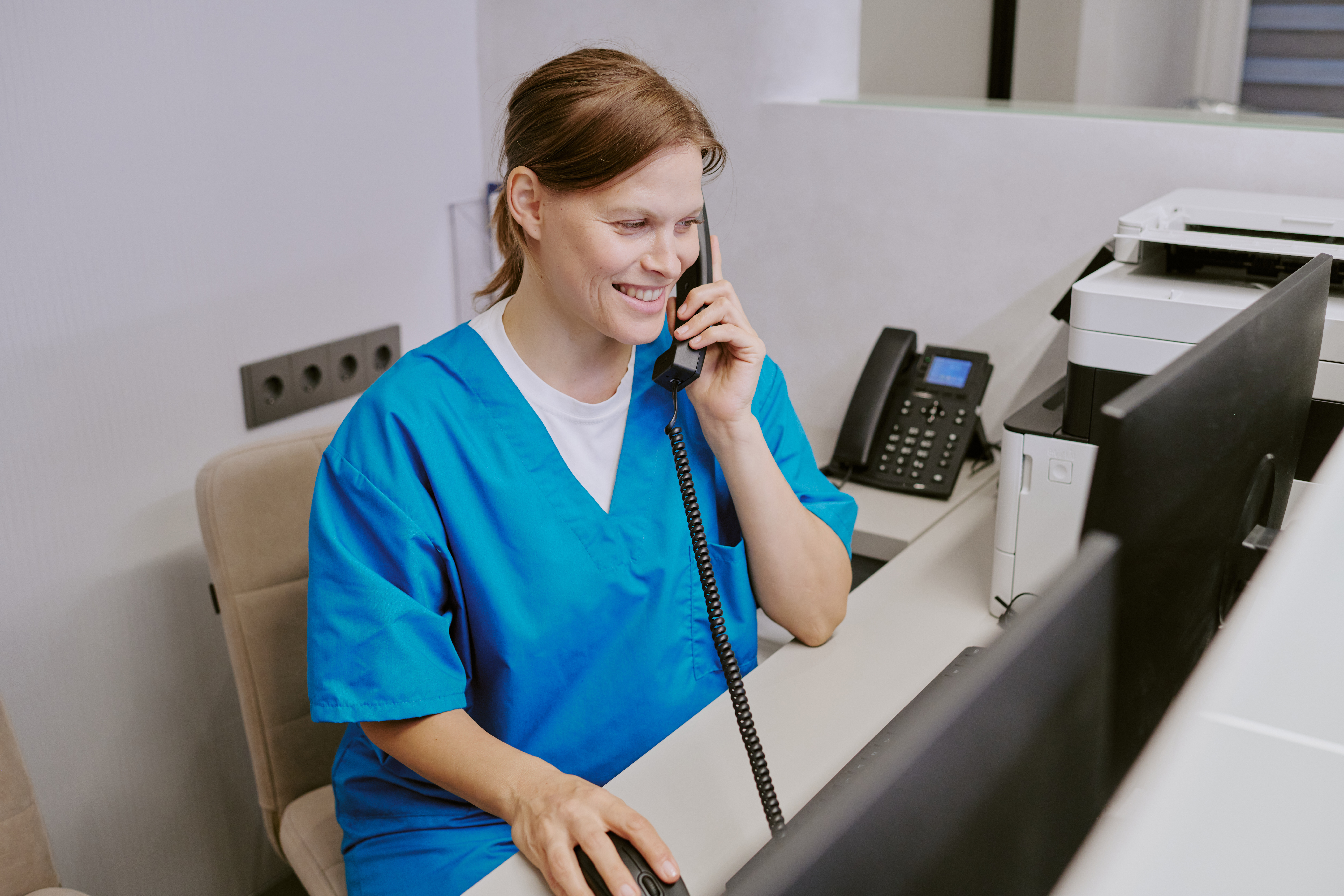 Young white woman with brown hair pulled back wearing blue scrubs answering a black landline phone at a medical reception desk