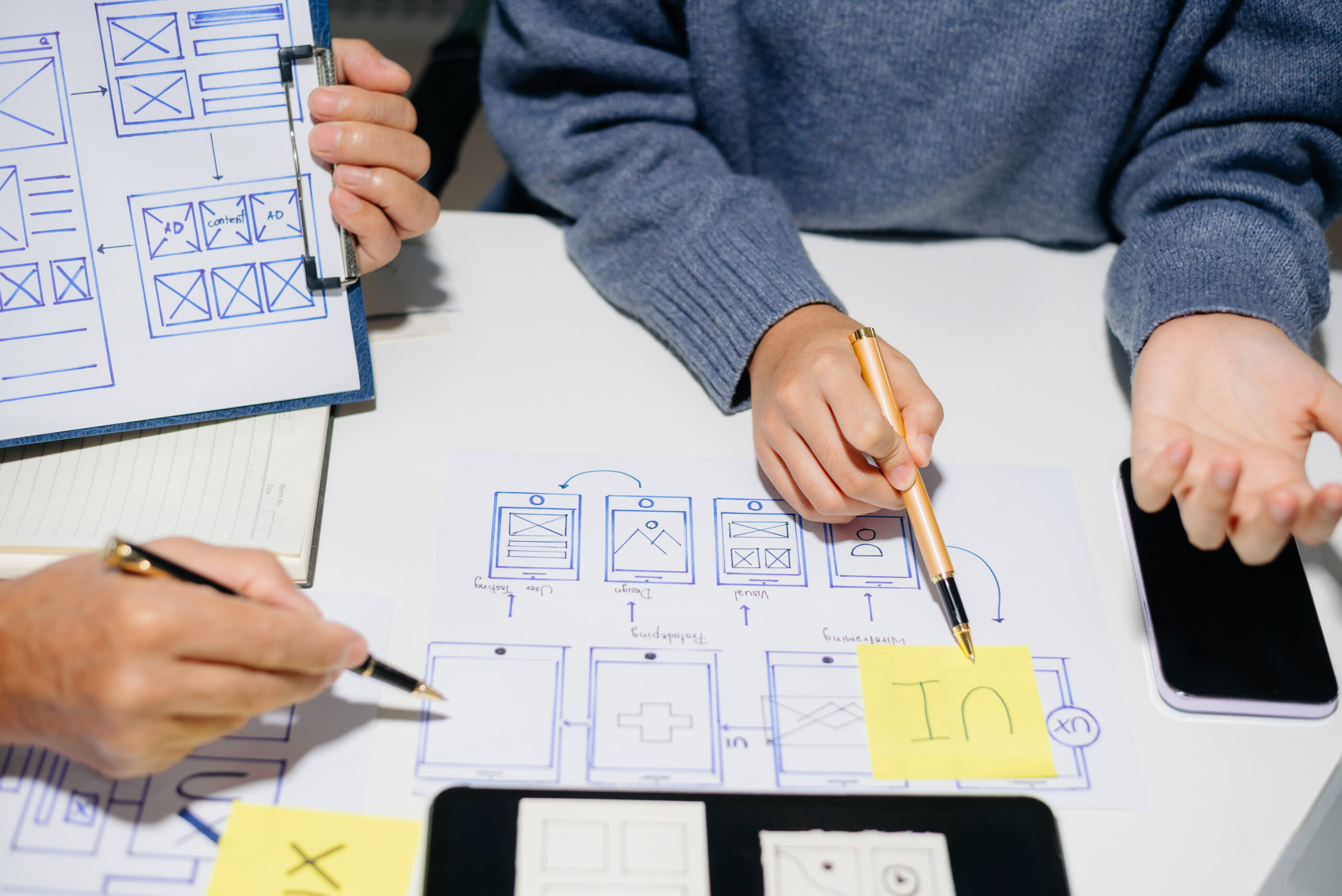 Close up of person's hands in a blue sweater using pens and digital toos to design blueprints