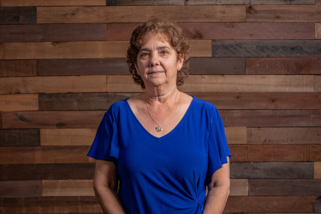 older woman with curly brown hair wearing a blue shirt and necklace in front of a wooden background